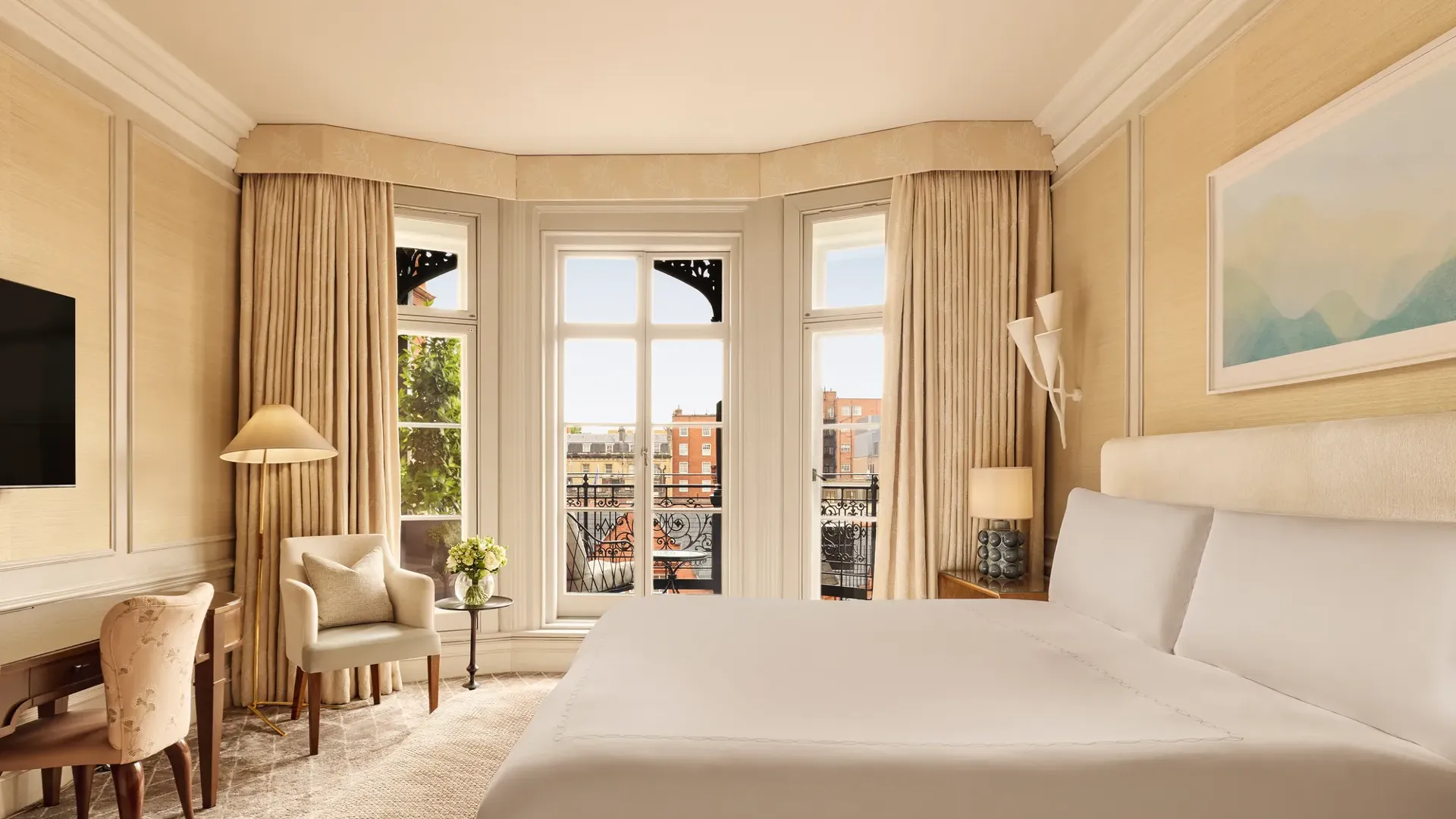 Bright bedroom with a large bed, writing desk, and armchair by tall bay windows draped in golden curtains, opening to a balcony with views of red-brick buildings.