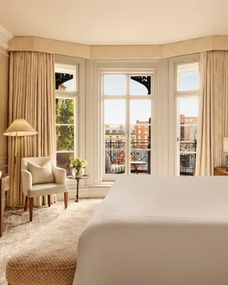 Bright bedroom with a large bed, writing desk, and armchair by tall bay windows draped in golden curtains, opening to a balcony with views of red-brick buildings.