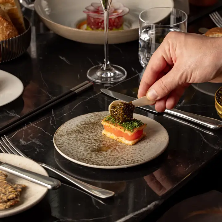 Hand topping a small salmon canapé with caviar at an elegantly set table with drinks and dishes.