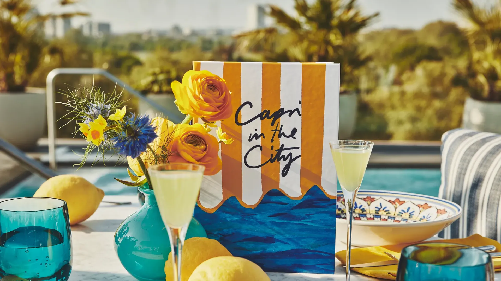 Poolside table with lemons, yellow cocktails, flowers, and a “Capri in the City” menu against palm trees.