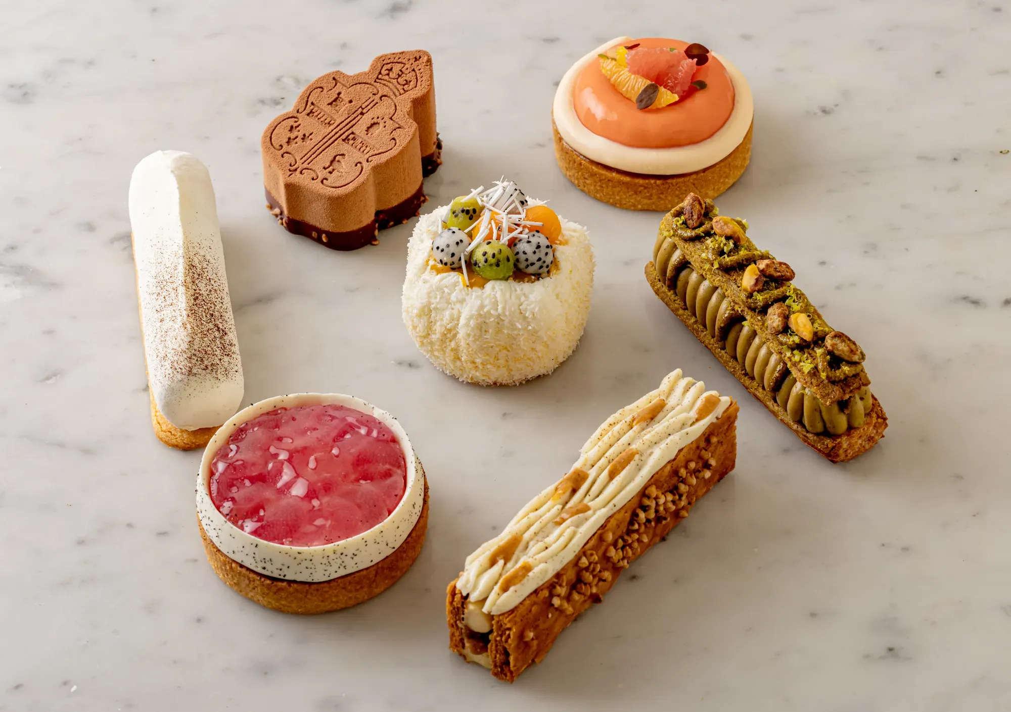 Assorted gourmet pastries on marble surface, including fruit tart, pistachio éclair, mousse cake, and citrus dessert with glaze