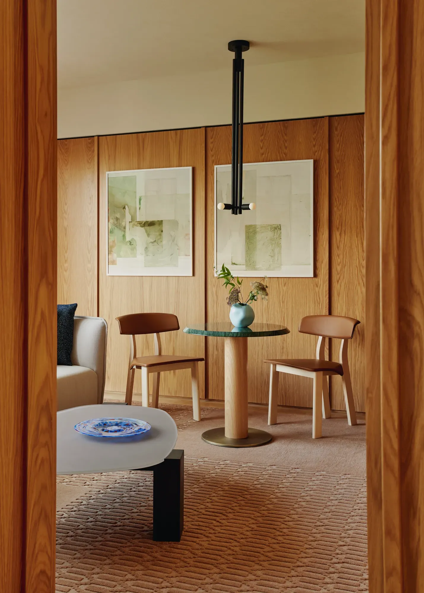 Small dining nook with green-topped table, wooden chairs, and soft artwork on wood-panelled walls with pendant light above.