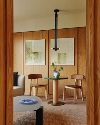 Small dining nook with green-topped table, wooden chairs, and soft artwork on wood-panelled walls with pendant light above.