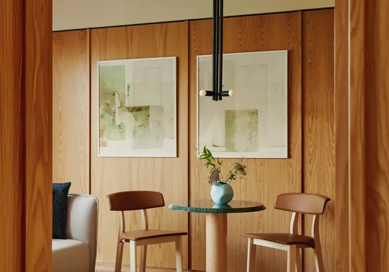 Small dining nook with green-topped table, wooden chairs, and soft artwork on wood-panelled walls with pendant light above.