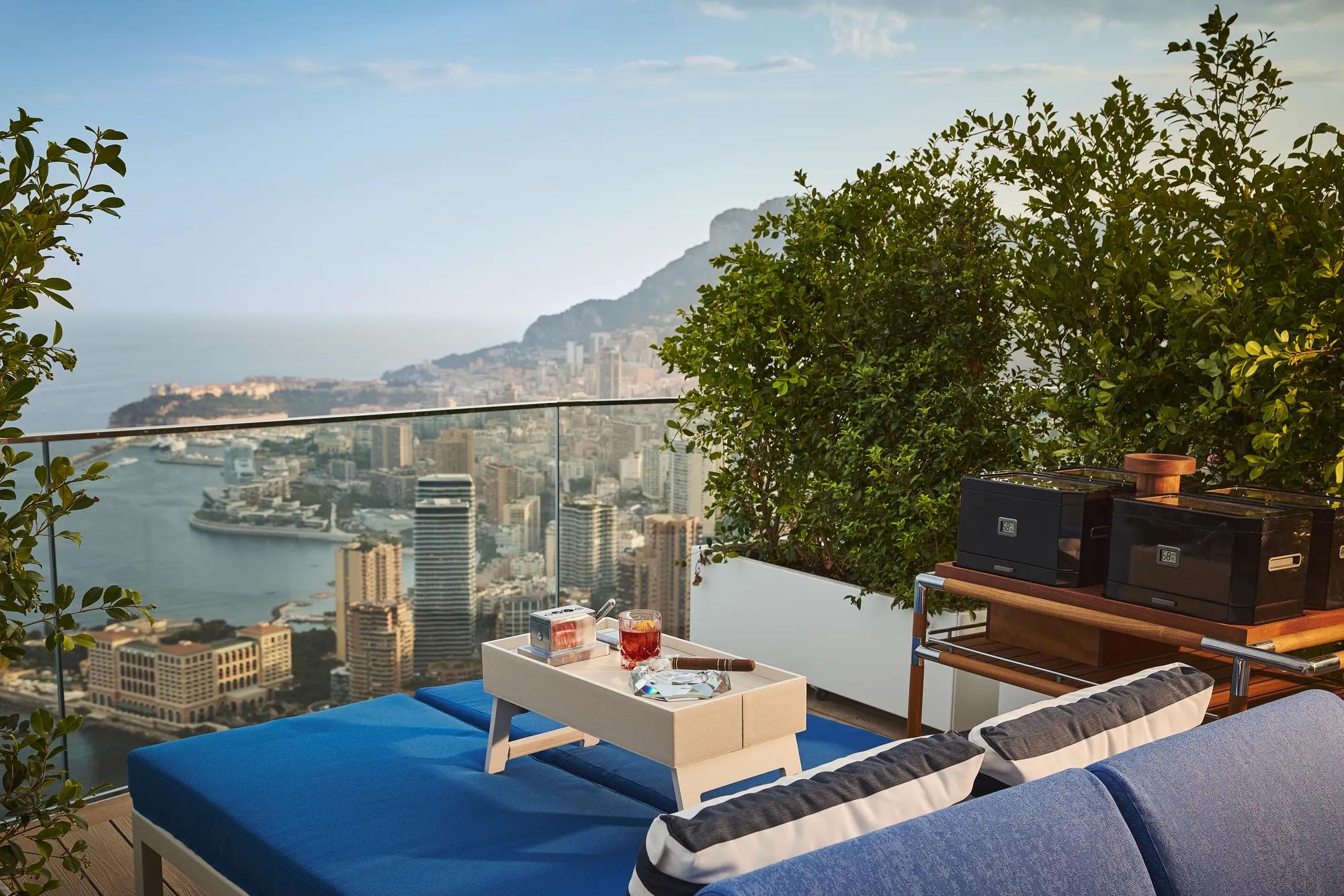 Rooftop terrace with blue sun loungers and a tray of drinks overlooking Monaco’s coastline, with buffet warmers placed nearby against lush greenery.