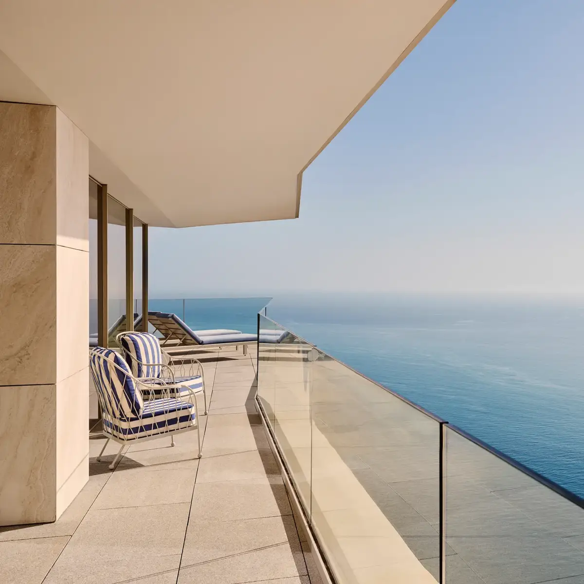 Balcony with striped chairs and loungers, glass balustrade, and uninterrupted sea view stretching beneath a clear blue sky.