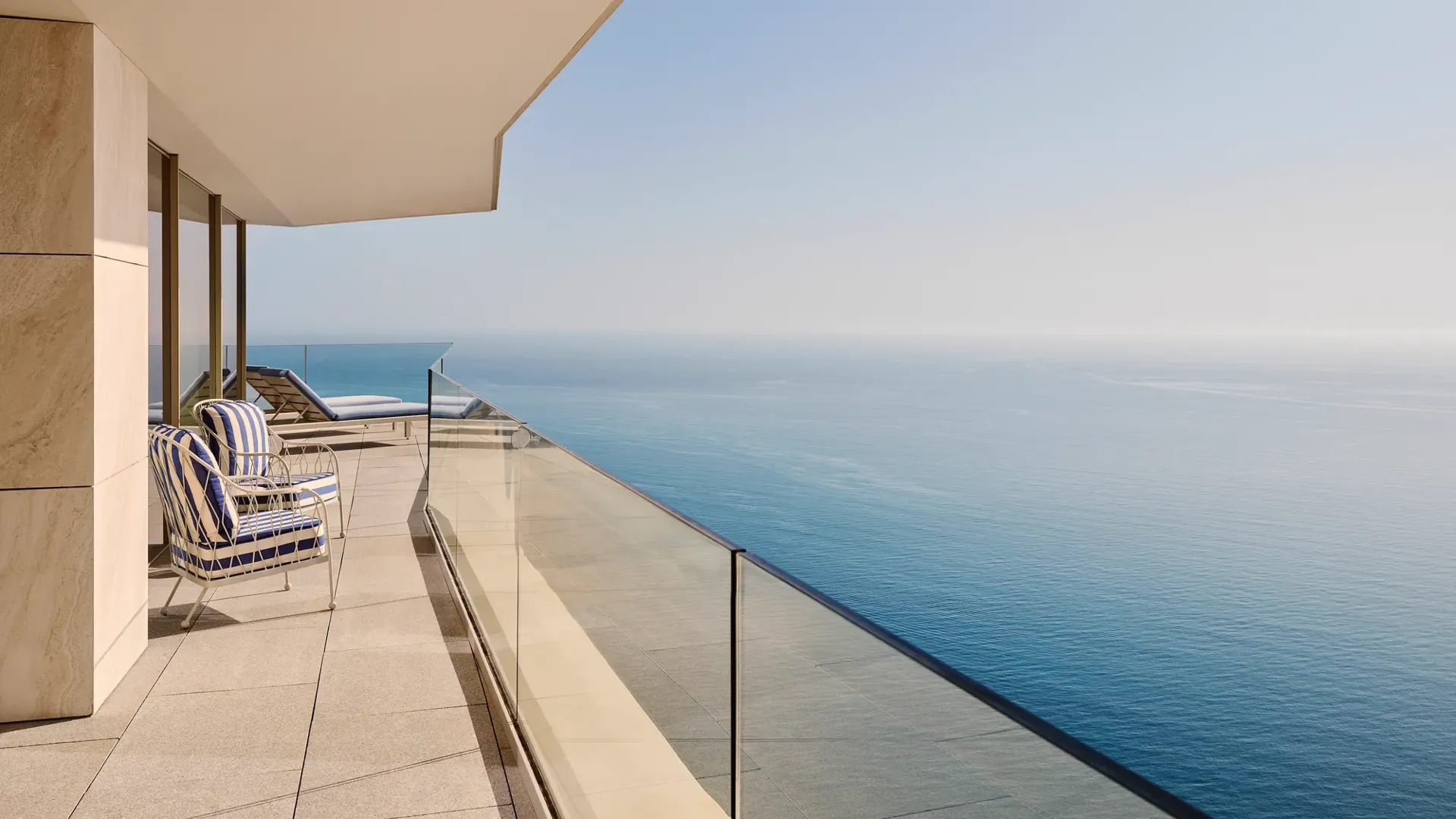 Balcon avec fauteuils rayés et transats, garde-corps en verre, et vue dégagée sur la mer sous un ciel bleu clair.