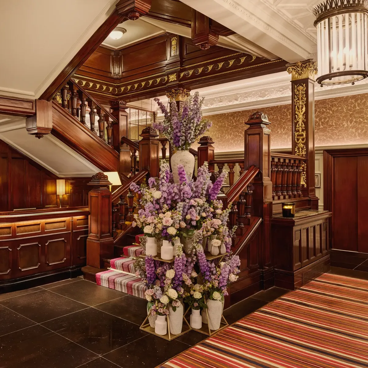Elegant hotel staircase with dark wood panelling and a large floral arrangement in soft purple and cream tones.