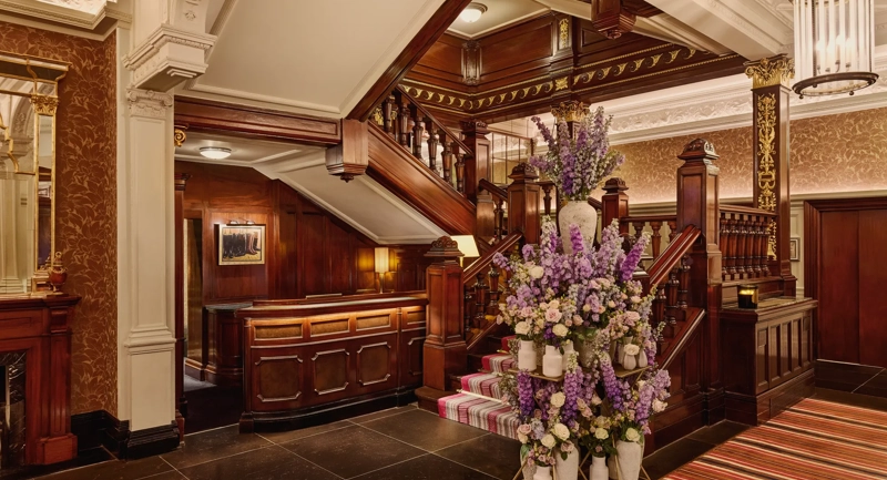Elegant hotel staircase with dark wood panelling and a large floral arrangement in soft purple and cream tones.