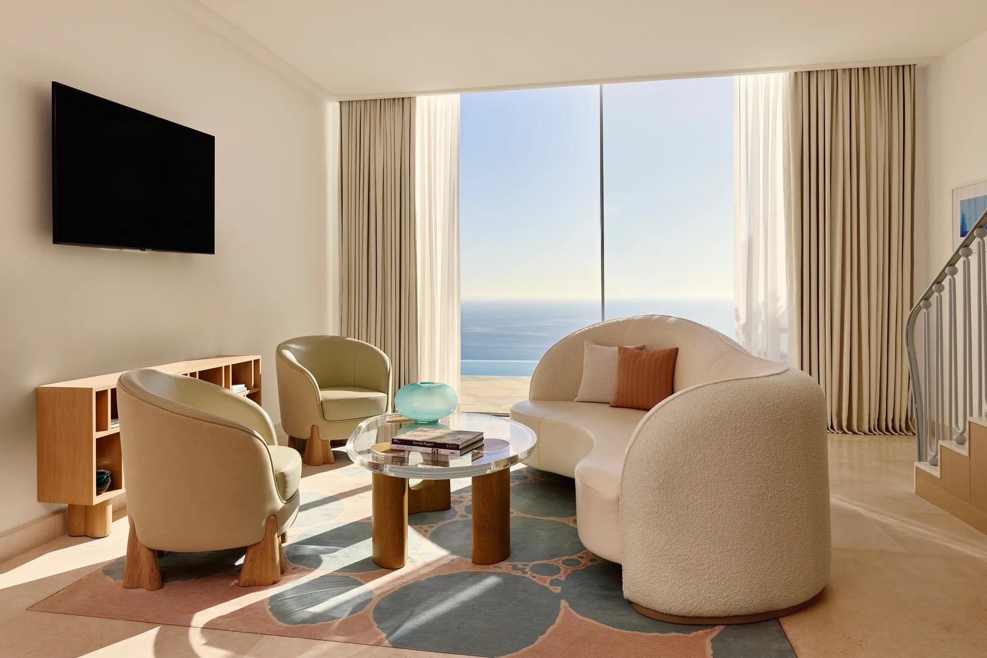 Salon lumineux avec canapé moderne incurvé, fauteuils et table basse en verre, donnant sur une vue panoramique de la mer.