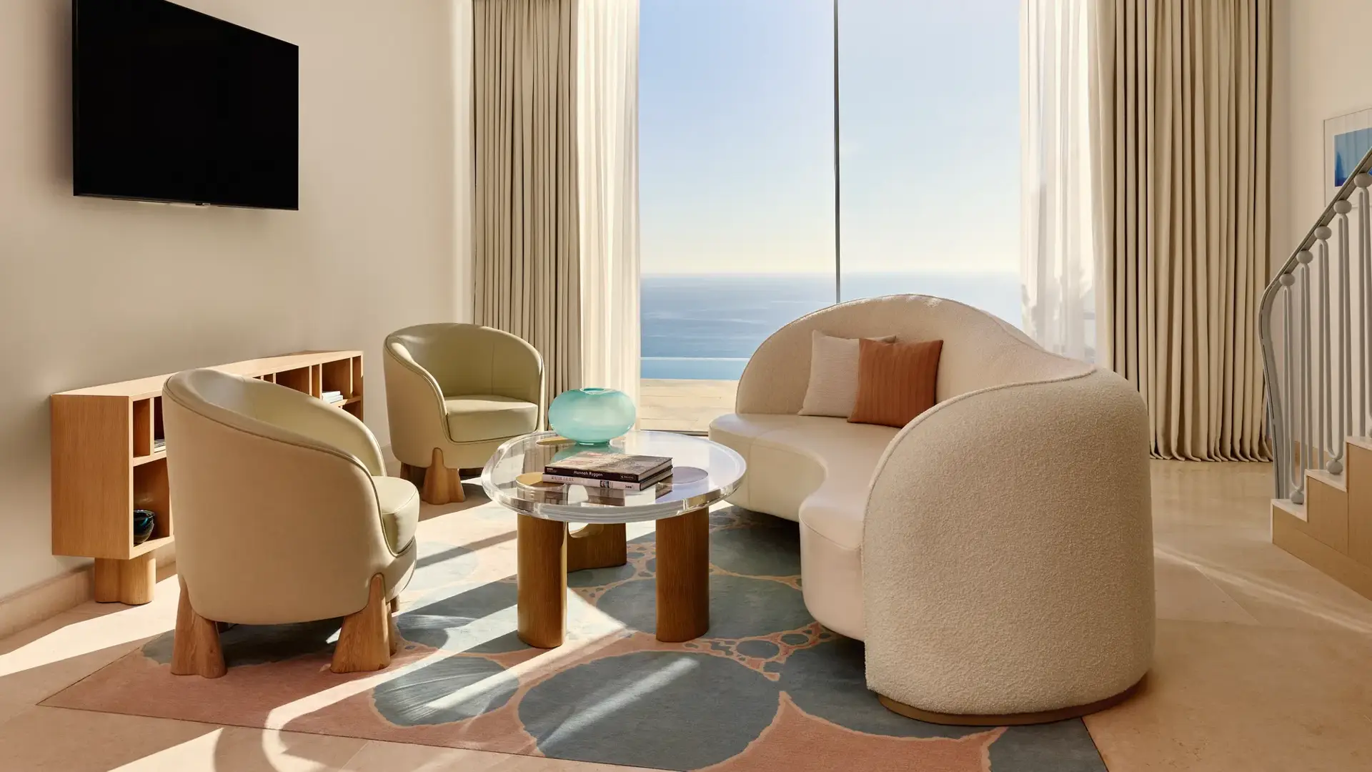 Salon lumineux avec canapé moderne incurvé, fauteuils et table basse en verre, donnant sur une vue panoramique de la mer.