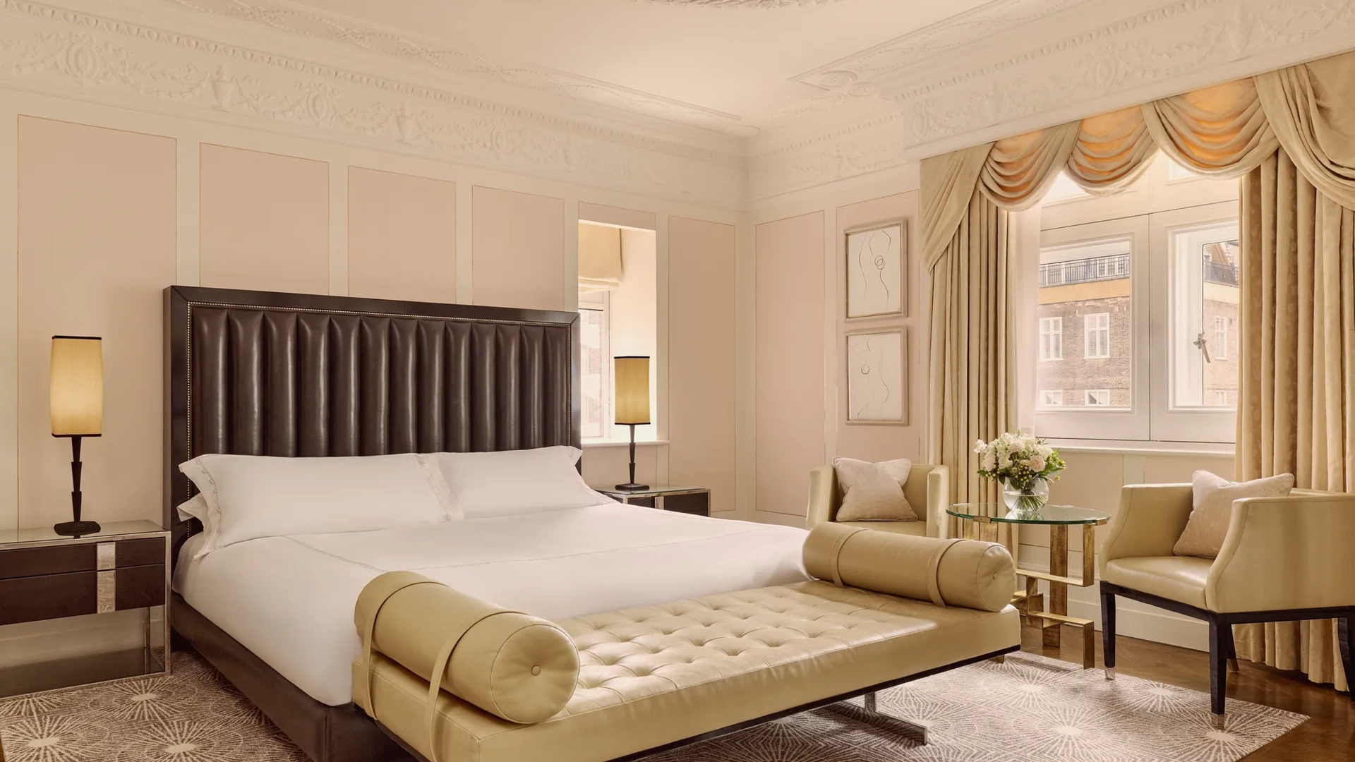 Elegant hotel bedroom with king bed, tufted bench, armchairs, and large window with drapes.