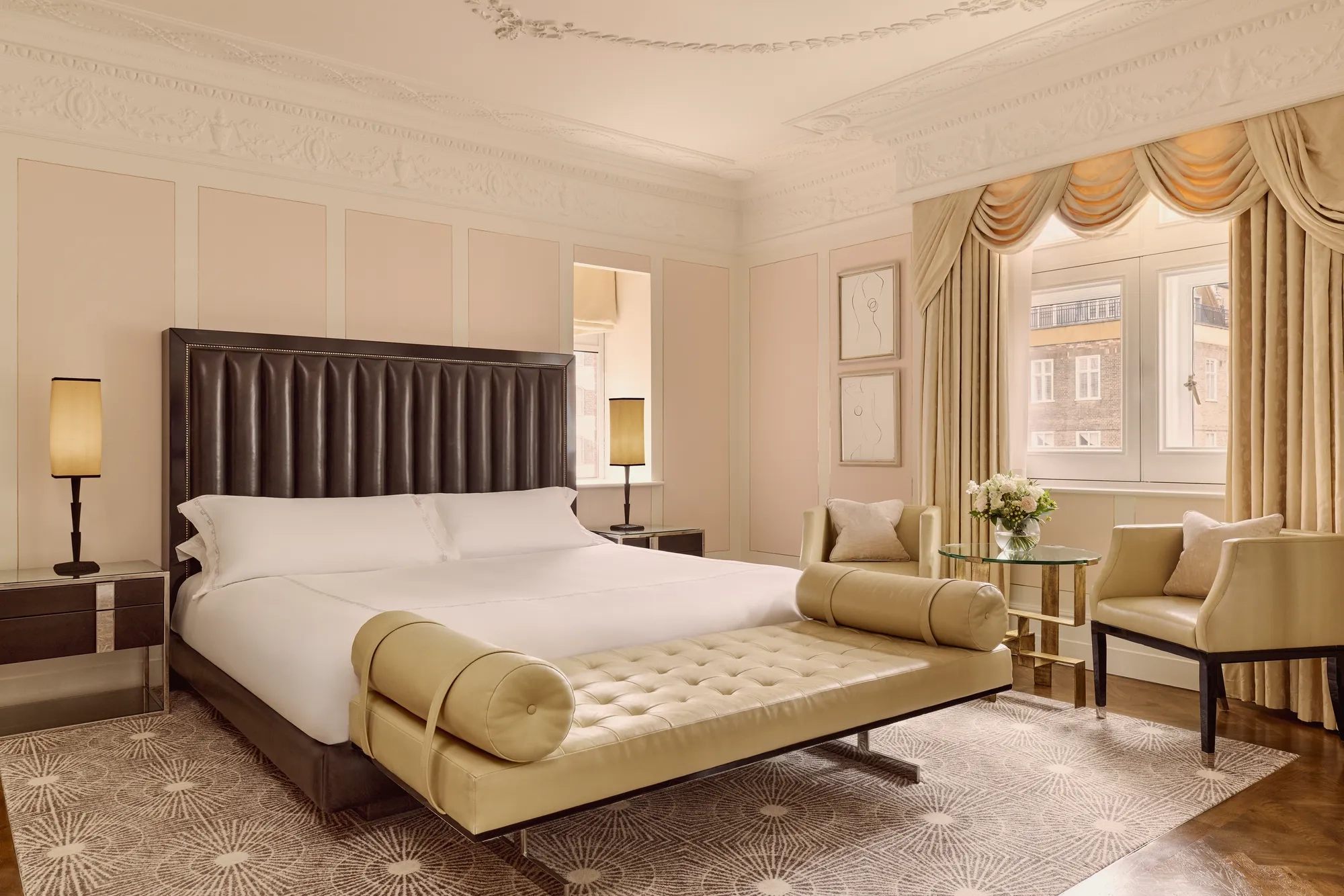 Elegant hotel bedroom with king bed, tufted bench, armchairs, and large window with drapes.