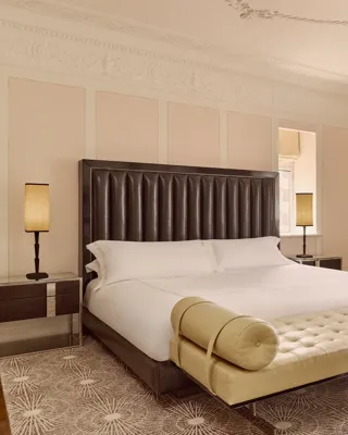 Elegant hotel bedroom with king bed, tufted bench, armchairs, and large window with drapes.