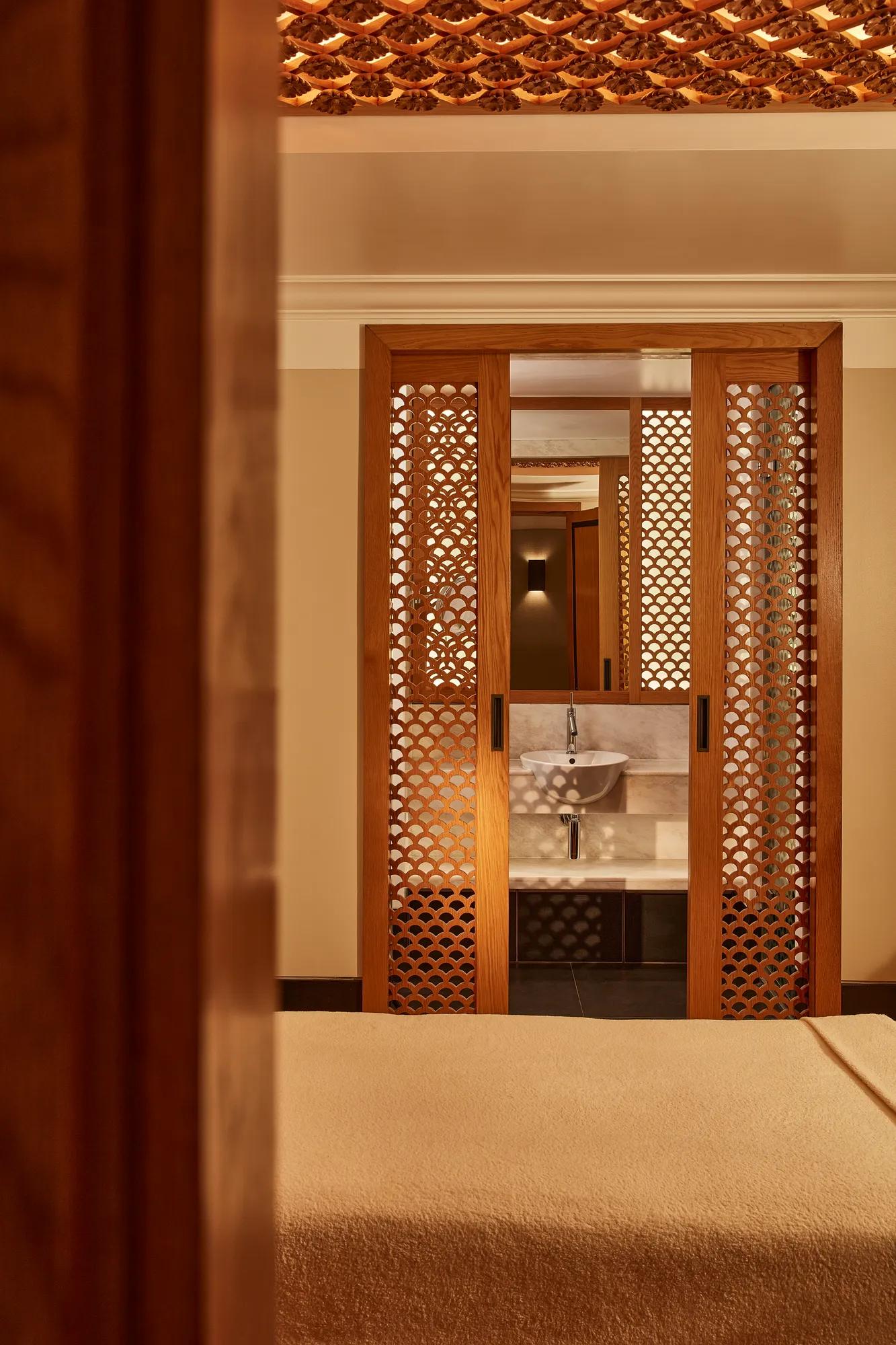 Peaceful spa room with a massage bed and wooden lattice doors revealing a small sink area in warm lighting.