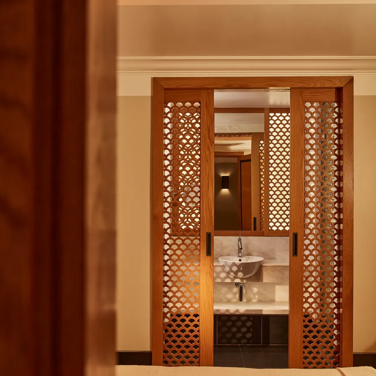 Peaceful spa room with a massage bed and wooden lattice doors revealing a small sink area in warm lighting.