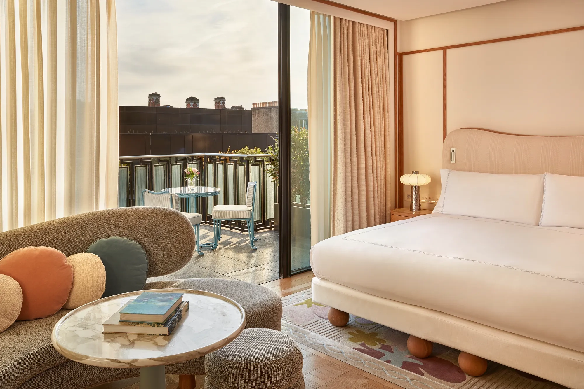 Contemporary bedroom with a curved sofa, marble coffee table, and king-size bed, opening onto a balcony with a bistro table and city views.