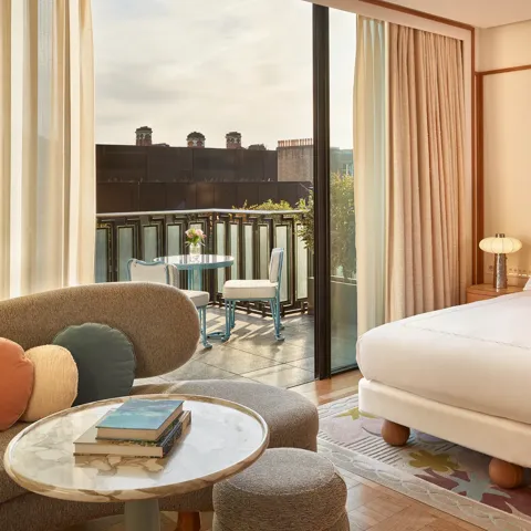 Contemporary bedroom with a curved sofa, marble coffee table, and king-size bed, opening onto a balcony with a bistro table and city views.