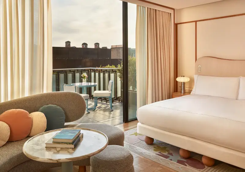 Contemporary bedroom with a curved sofa, marble coffee table, and king-size bed, opening onto a balcony with a bistro table and city views.