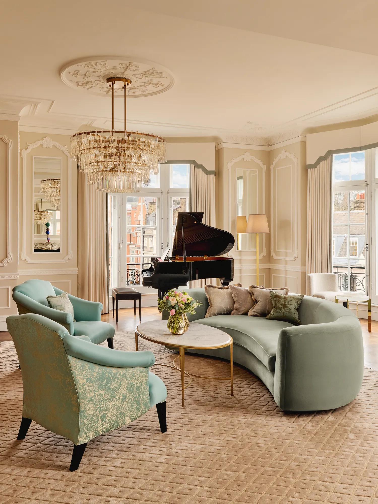 Elegant living room with a curved mint-green sofa, two patterned armchairs, and a round marble coffee table under a crystal chandelier, with a grand piano by tall windows framed with cream drapes.