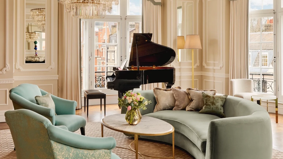Elegant living room with a curved mint-green sofa, two patterned armchairs, and a round marble coffee table under a crystal chandelier, with a grand piano by tall windows framed with cream drapes.