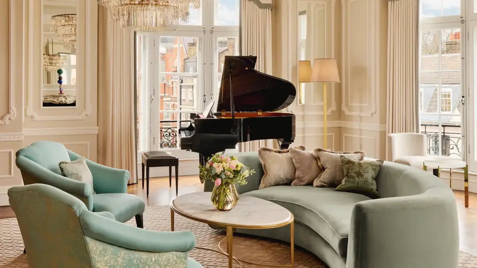 Elegant living room with a curved mint-green sofa, two patterned armchairs, and a round marble coffee table under a crystal chandelier, with a grand piano by tall windows framed with cream drapes.