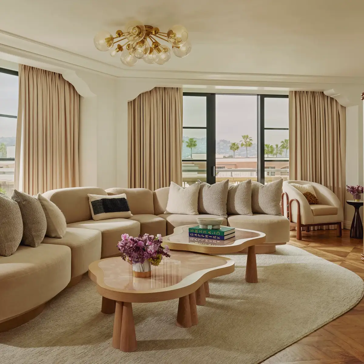 California Suite living room with curved beige sectional, bubble‑glass chandelier, twin pink stone coffee tables, floor‑to‑ceiling windows with drapes, and a view of palms.