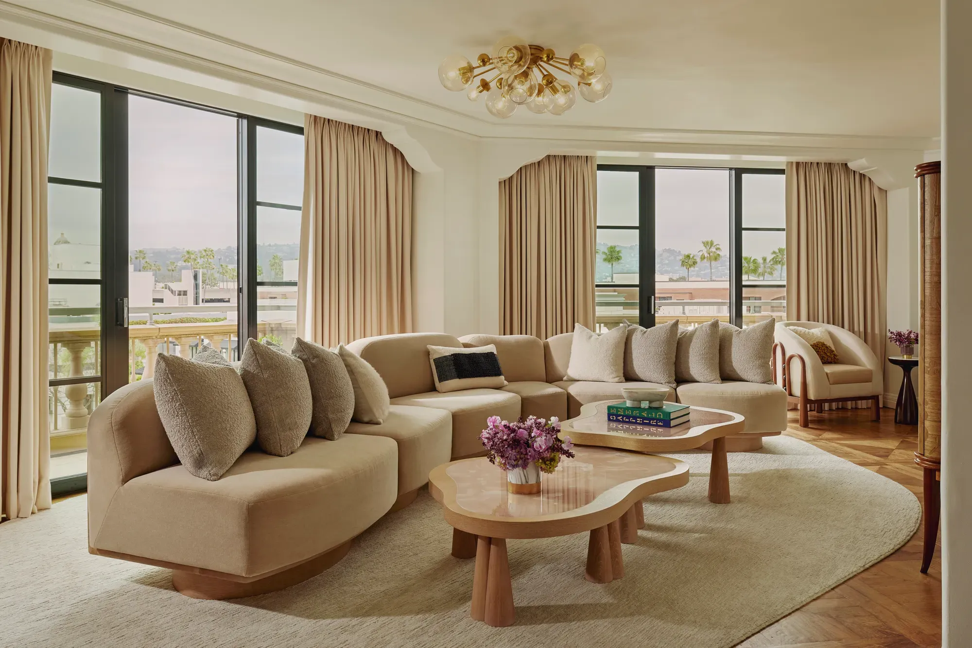 California Suite living room with curved beige sectional, bubble‑glass chandelier, twin pink stone coffee tables, floor‑to‑ceiling windows with drapes, and a view of palms.