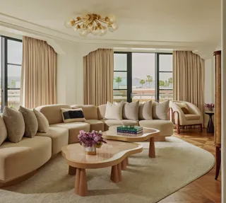 California Suite living room with curved beige sectional, bubble‑glass chandelier, twin pink stone coffee tables, floor‑to‑ceiling windows with drapes, and a view of palms.
