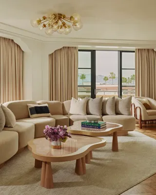California Suite living room with curved beige sectional, bubble‑glass chandelier, twin pink stone coffee tables, floor‑to‑ceiling windows with drapes, and a view of palms.