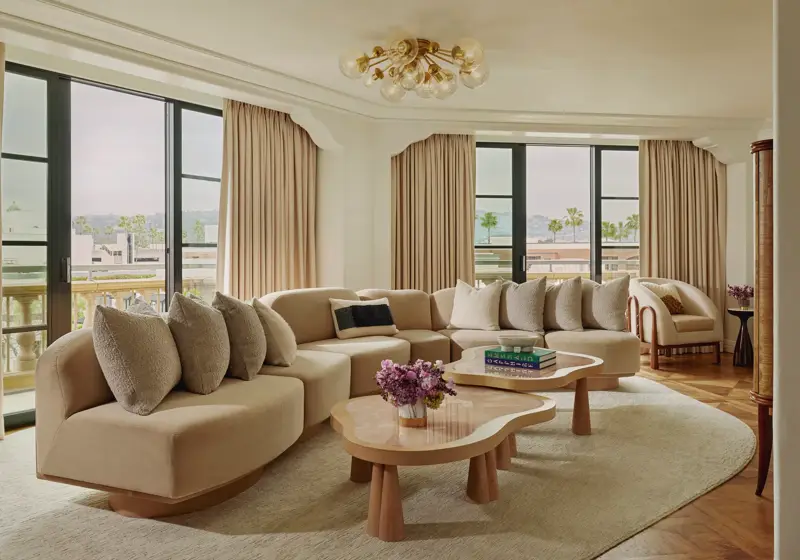 California Suite living room with curved beige sectional, bubble‑glass chandelier, twin pink stone coffee tables, floor‑to‑ceiling windows with drapes, and a view of palms.