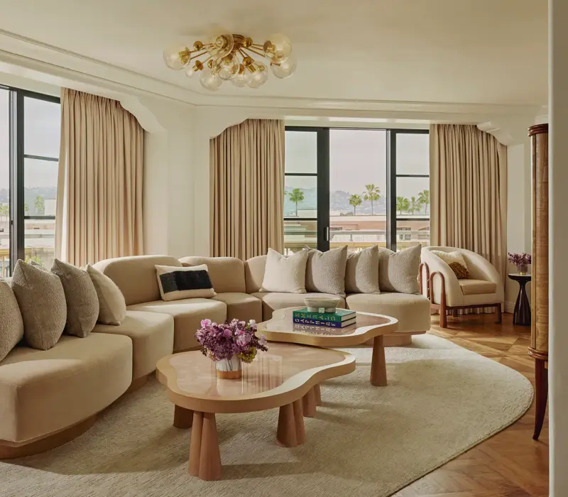 California Suite living room with curved beige sectional, bubble‑glass chandelier, twin pink stone coffee tables, floor‑to‑ceiling windows with drapes, and a view of palms.