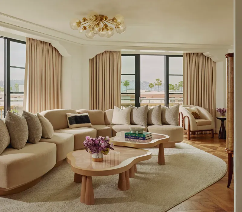 California Suite living room with curved beige sectional, bubble‑glass chandelier, twin pink stone coffee tables, floor‑to‑ceiling windows with drapes, and a view of palms.
