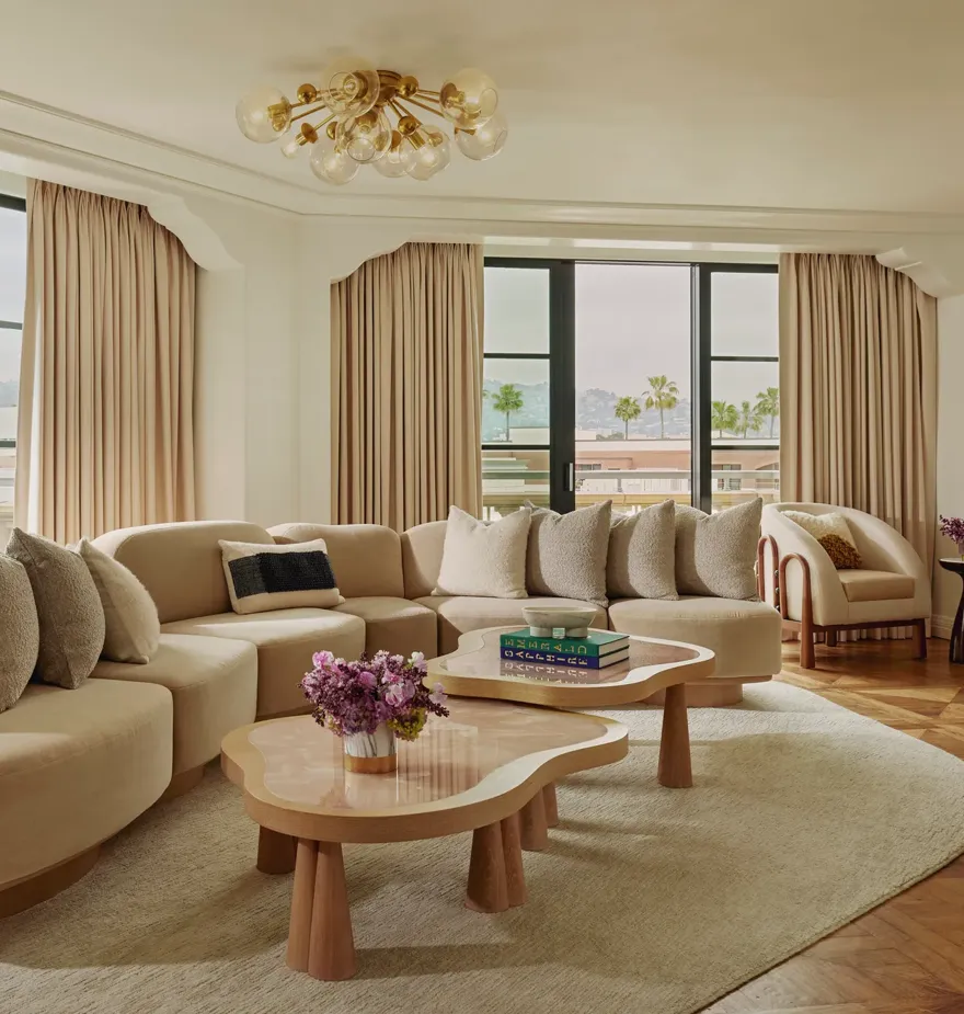 California Suite living room with curved beige sectional, bubble‑glass chandelier, twin pink stone coffee tables, floor‑to‑ceiling windows with drapes, and a view of palms.
