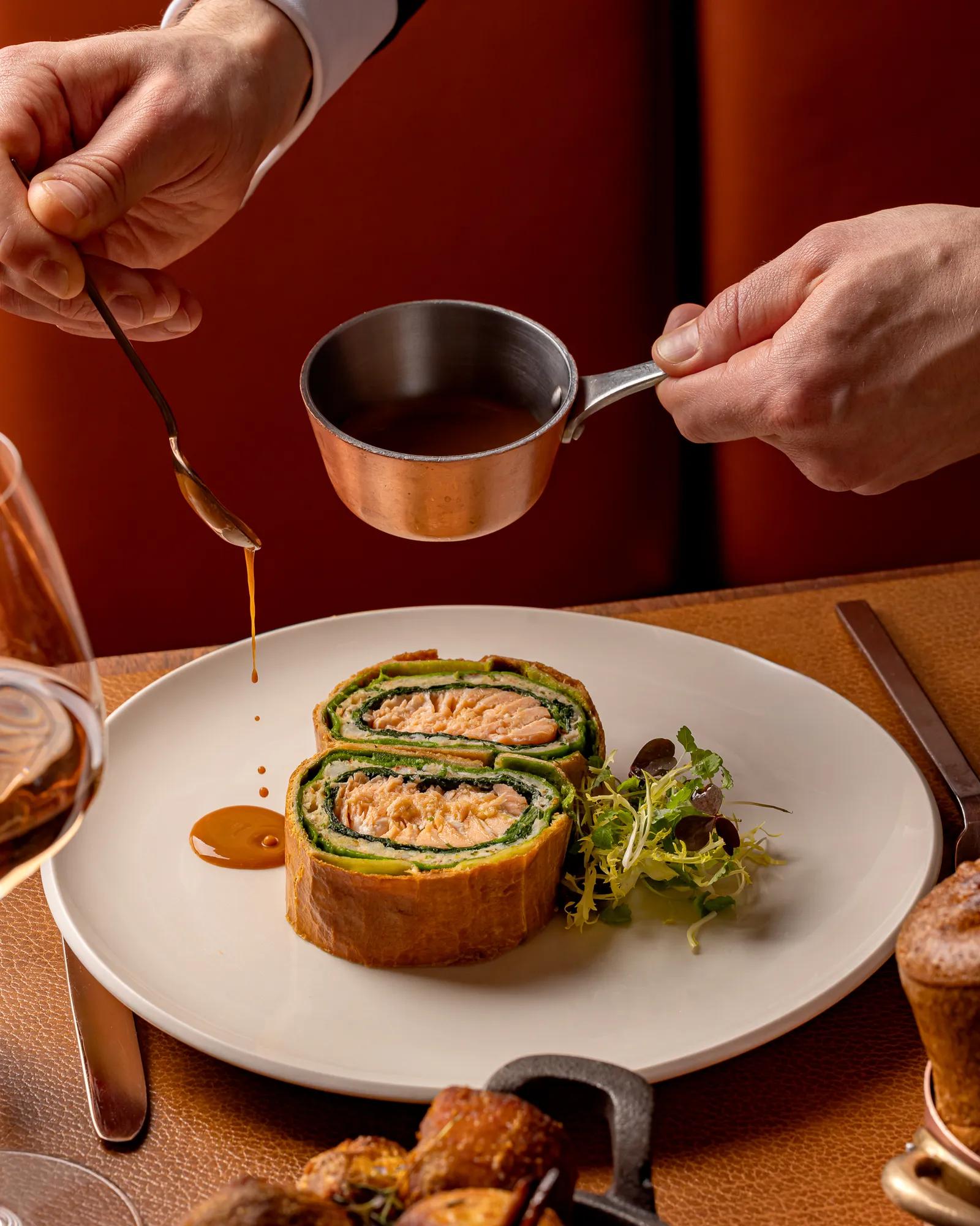 Sauce being poured onto a plated salmon Wellington served with fresh greens at a fine dining table