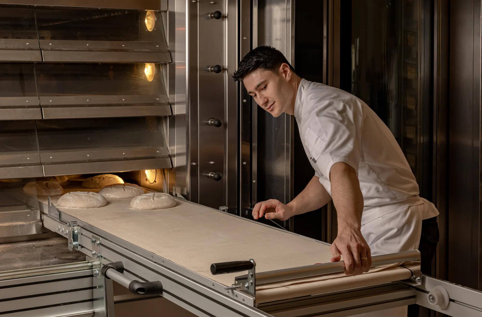 Chef in white uniform monitoring dough on conveyor entering oven in commercial bakery kitchen.