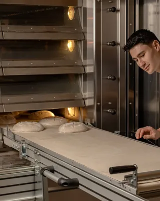 Chef in white uniform monitoring dough on conveyor entering oven in commercial bakery kitchen.