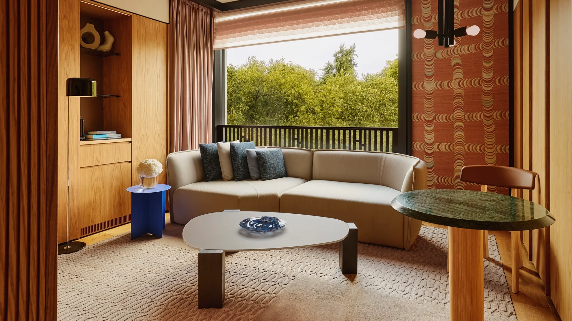 Modern living room with a neutral sectional sofa, blue and gray pillows, large window overlooking green trees.