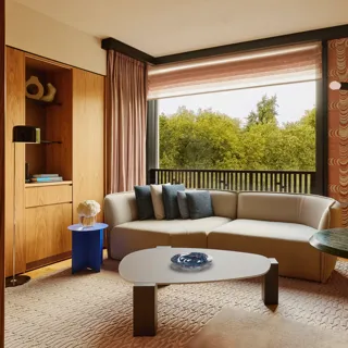 Modern living room with a neutral sectional sofa, blue and gray pillows, large window overlooking green trees.