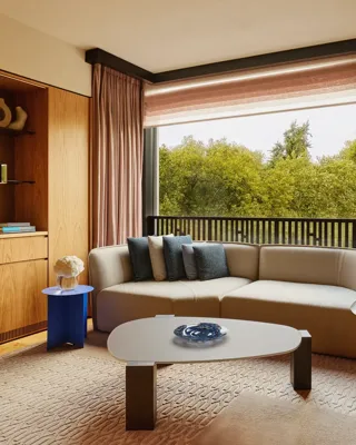 Modern living room with a neutral sectional sofa, blue and gray pillows, large window overlooking green trees.