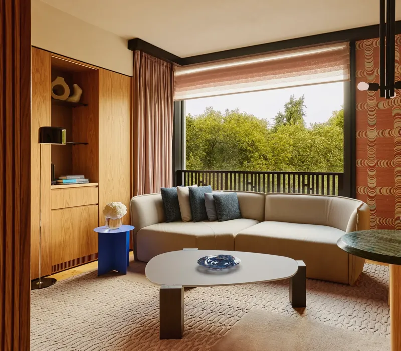 Modern living room with a neutral sectional sofa, blue and gray pillows, large window overlooking green trees.