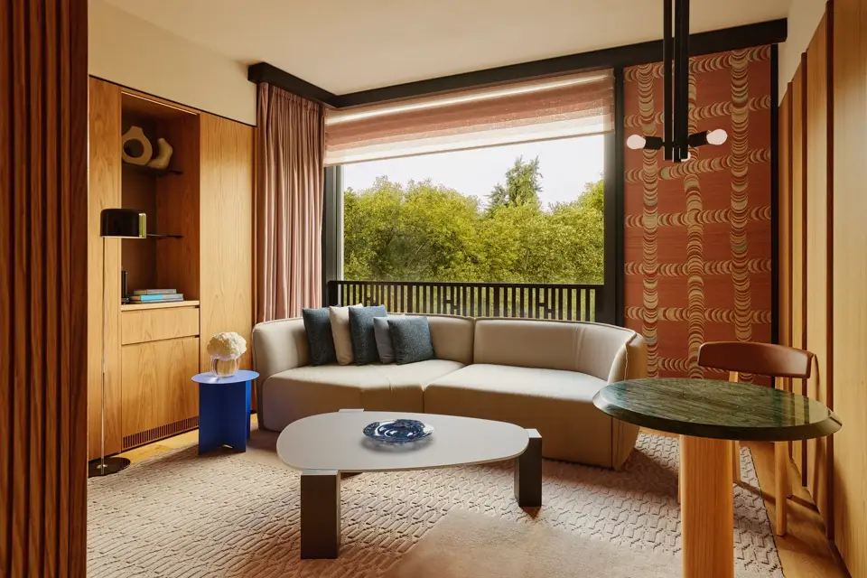 Modern living room with a neutral sectional sofa, blue and gray pillows, large window overlooking green trees.