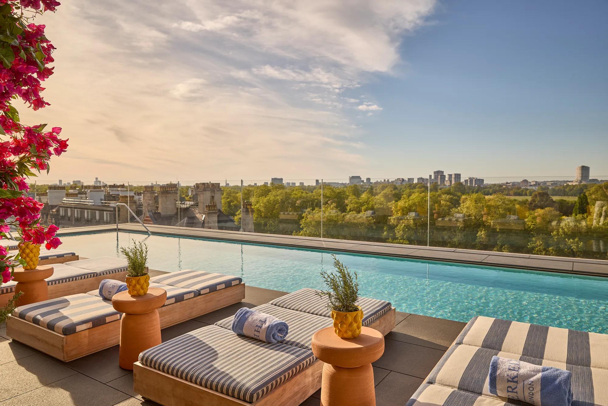 Poolside loungers with striped cushions beside a rooftop infinity pool overlooking treetops and a city skyline at sunset.