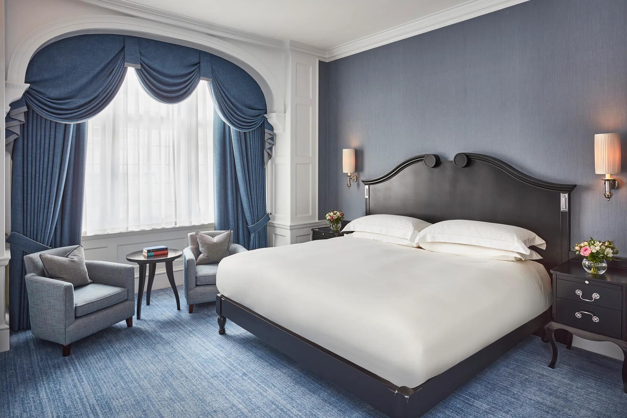Elegant bedroom with a dark wood bed, blue upholstered chairs, and arched window draped in blue curtains.