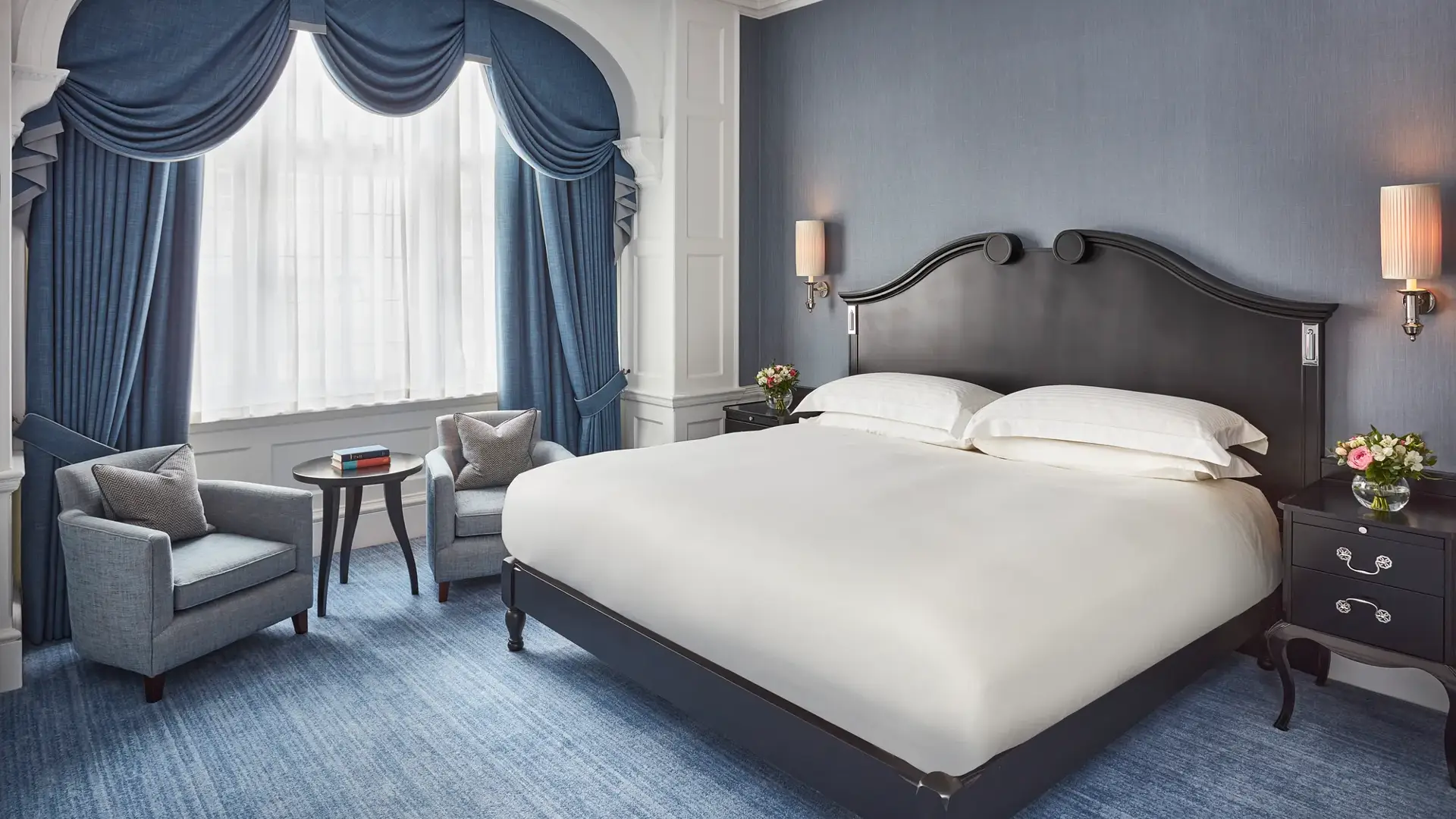 Elegant bedroom with a dark wood bed, blue upholstered chairs, and arched window draped in blue curtains.