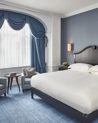 Elegant bedroom with a dark wood bed, blue upholstered chairs, and arched window draped in blue curtains.
