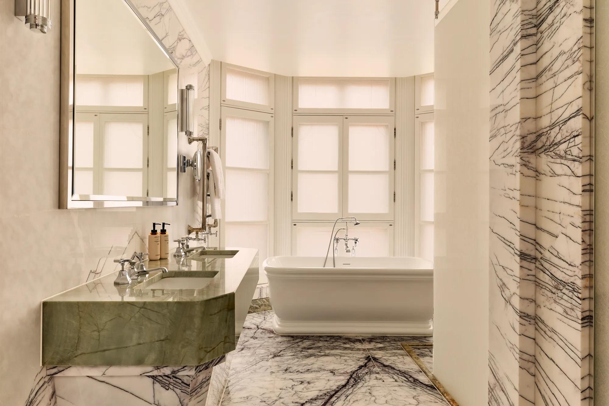 Luxurious marble bathroom featuring a freestanding white bathtub in front of frosted bay windows, with a double green marble vanity and large wall mirror on the left.
