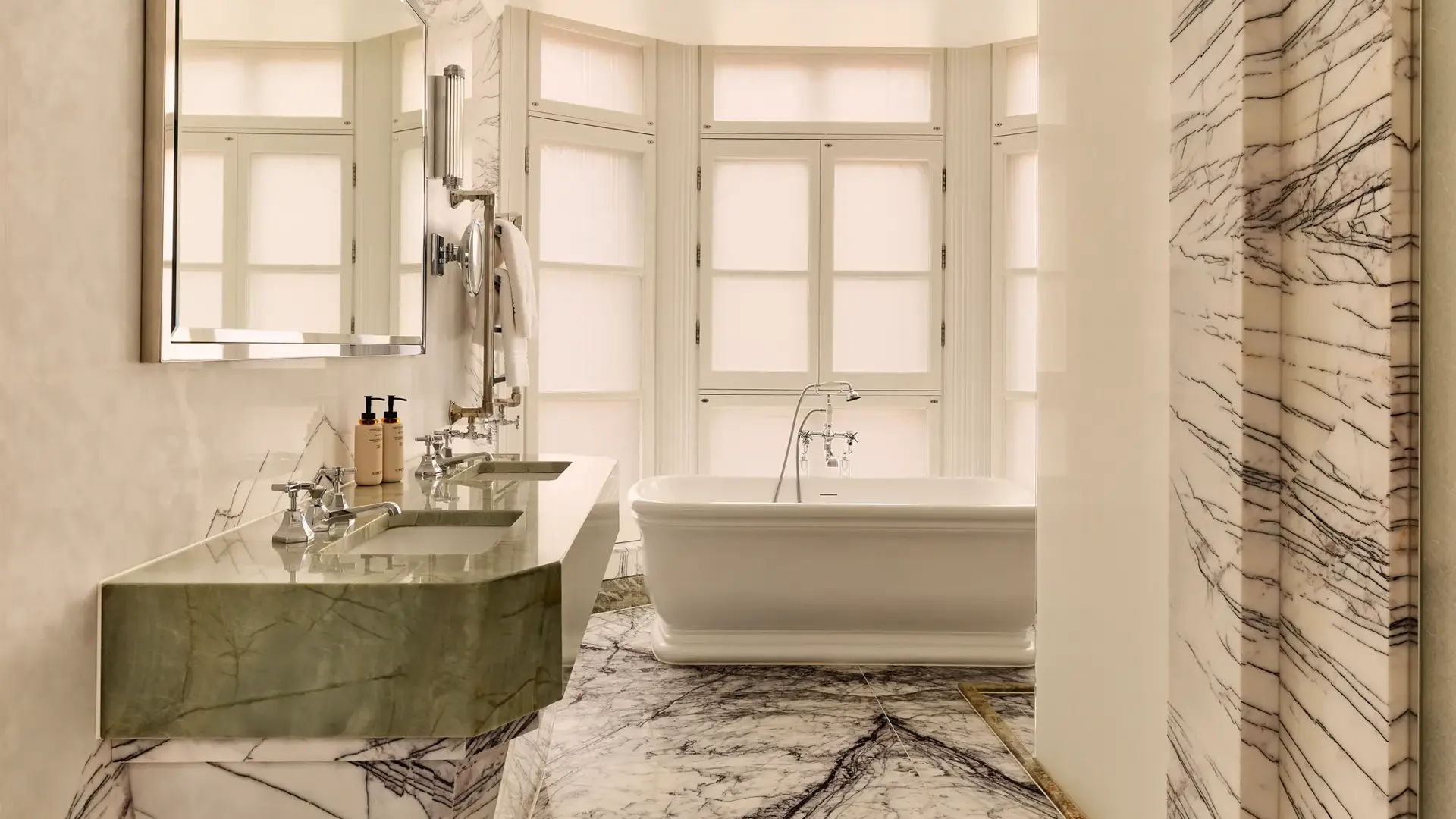 Luxurious marble bathroom featuring a freestanding white bathtub in front of frosted bay windows, with a double green marble vanity and large wall mirror on the left.