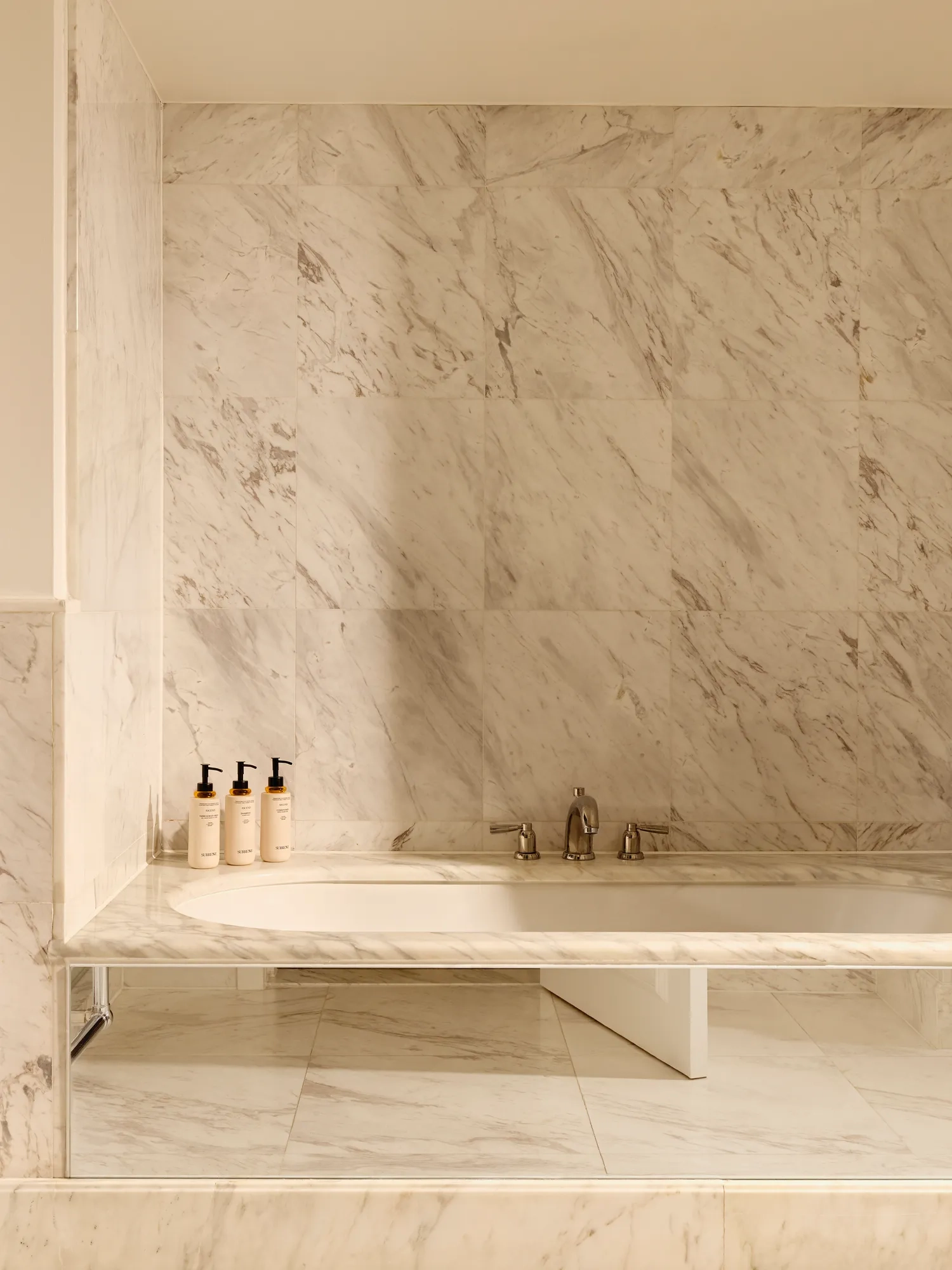 Bathroom in white marble with a built-in bathtub, chrome fixtures, and luxury toiletries displayed on the ledge.