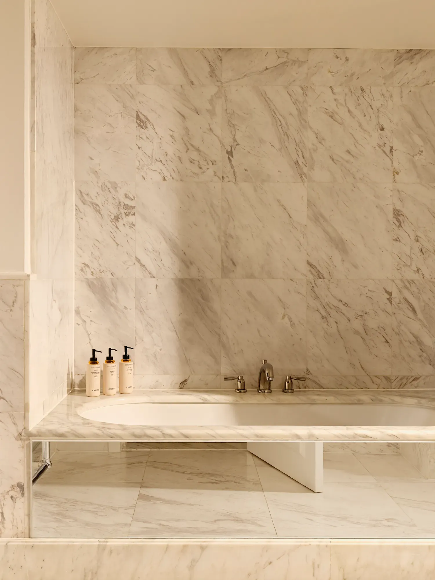  Bathroom in white marble with a built-in bathtub, chrome fixtures, and luxury toiletries displayed on the ledge.